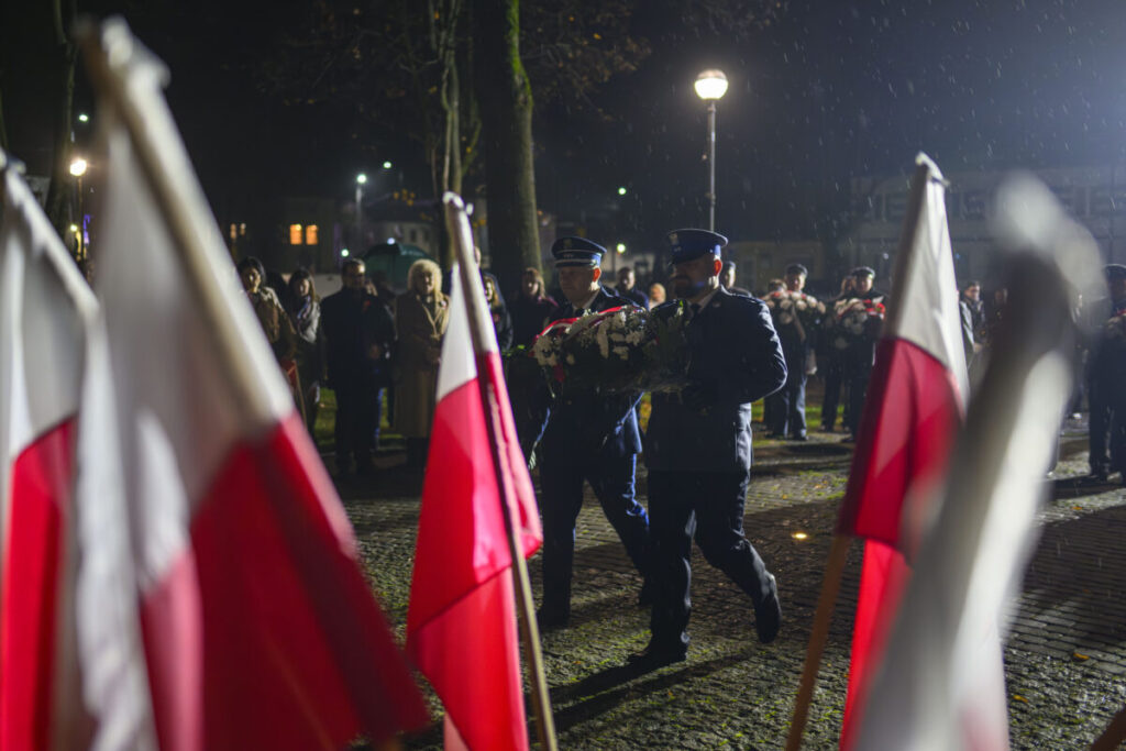 Dwóch mężczyzn w mundurach niesie wieniec podczas nocnej uroczystości. Otaczają ich polskie flagi. W tle widoczne są postacie i latarnia uliczna.