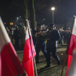 Dwóch mężczyzn w mundurach niesie wieniec podczas nocnej uroczystości. Otaczają ich polskie flagi. W tle widoczne są postacie i latarnia uliczna.