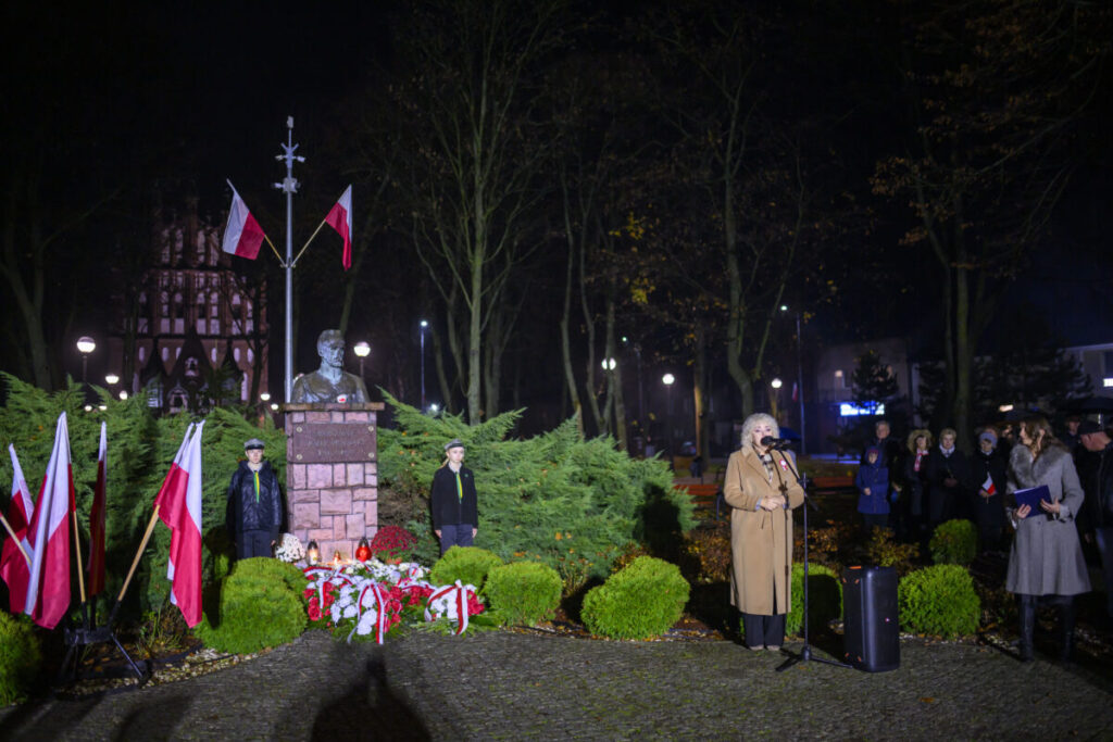 Pomnik z popiersiem otoczony flagami Polski, wieńce kwiatowe z biało-czerwonymi wstęgami. W tle ciemny park, z przodu kobieta przy mikrofonie. Obok młodzieńcze harcerze w mundurach.