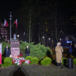 Pomnik z popiersiem otoczony flagami Polski, wieńce kwiatowe z biało-czerwonymi wstęgami. W tle ciemny park, z przodu kobieta przy mikrofonie. Obok młodzieńcze harcerze w mundurach.