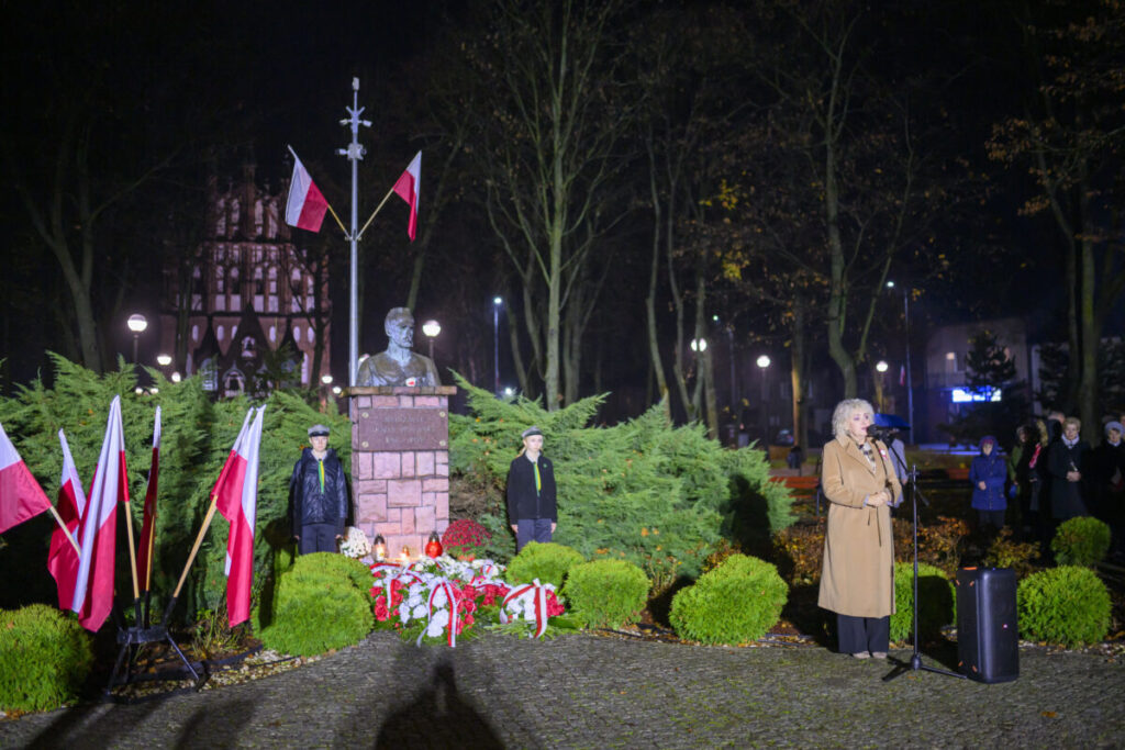 Pomnik z popiersiem otoczony zielenią, udekorowany biało-czerwonymi flagami i kwiatami. Obok kobieta przemawia do mikrofonu. Nocne oświetlenie, w tle drzewa i budynek.