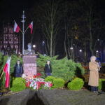 Pomnik z popiersiem otoczony zielenią, udekorowany biało-czerwonymi flagami i kwiatami. Obok kobieta przemawia do mikrofonu. Nocne oświetlenie, w tle drzewa i budynek.