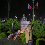 Osoba w płaszczu przemawia przy mikrofonie przed pomnikiem, w tle flagi Polski. Zieleń roślin i nocne oświetlenie. Bukiety kwiatów u podstawy.