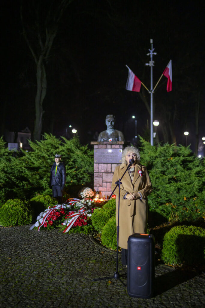 Osoba w płaszczu przemawia przy mikrofonie przed pomnikiem, w tle flagi Polski. Zieleń roślin i nocne oświetlenie. Bukiety kwiatów u podstawy.