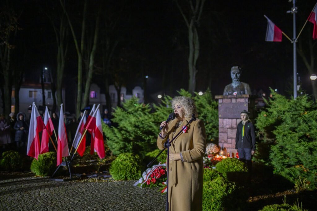 Kobieta w płaszczu przemawia przed mikrofonem obok flag Polski. Tło to nocny park z pomnikiem i zielonymi krzewami. Widać także znicze i osoby w mundurach.