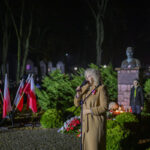 Kobieta w płaszczu przemawia przed mikrofonem obok flag Polski. Tło to nocny park z pomnikiem i zielonymi krzewami. Widać także znicze i osoby w mundurach.