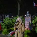 Osoba przemawia nocą na tle pomnika i zielonych krzewów. Ubrana w beżowy płaszcz, trzyma mikrofon. W tle widać biało-czerwoną flagę.