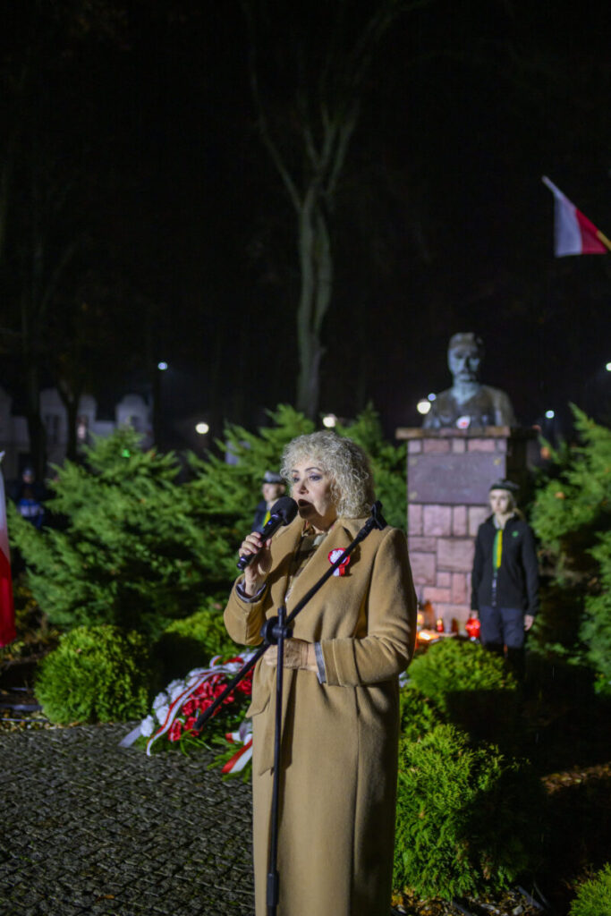 Osoba przemawia nocą na tle pomnika i zielonych krzewów. Ubrana w beżowy płaszcz, trzyma mikrofon. W tle widać biało-czerwoną flagę.