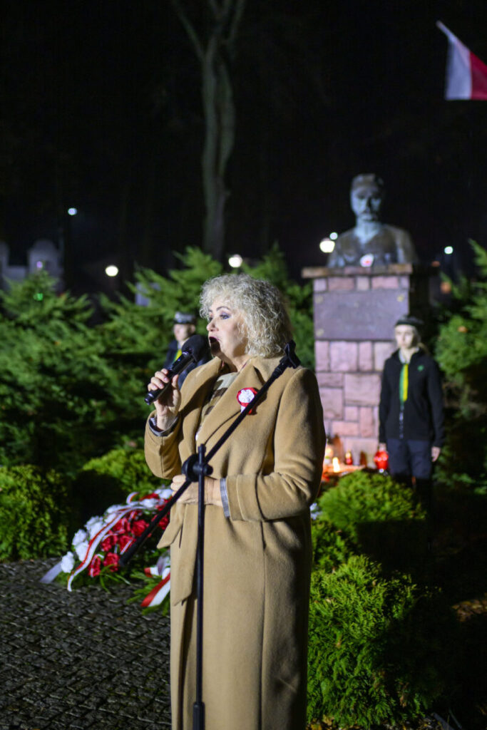 Kobieta w beżowym płaszczu mówi do mikrofonu przed pomnikiem na tle zielonych krzewów. W tle noc, zapalone znicze i flaga. Obok stoi młoda osoba w mundurku.