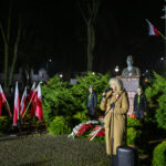 Kobieta w płaszczu przemawia przy mikrofonie w parku. W tle są flagi polskie i pomnik. Nocna sceneria, kolorowe światło.