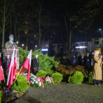 Na zdjęciu jest popiersie przyozdobione flagami Polski w zielonym parku. Kobieta w płaszczu przemawia do mikrofonu. W tle grupa ludzi stoi nocą.