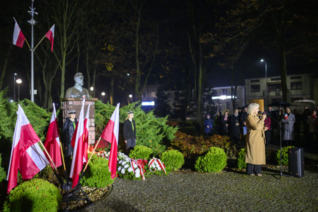 Pomnik otoczony flagami Polski, nocą, z przemawiającą osobą po prawej. Tło stanowią drzewa i zgromadzony tłum.