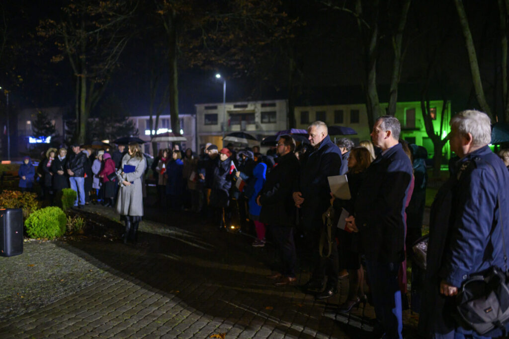 Grupa ludzi w parku po zmroku, ubranych w ciemne płaszcze, otoczona drzewami. W tle budynki z zielonym oświetleniem.
