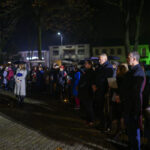 Grupa ludzi w parku po zmroku, ubranych w ciemne płaszcze, otoczona drzewami. W tle budynki z zielonym oświetleniem.