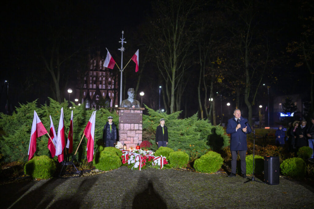 Mężczyzna przemawia podczas uroczystości przy pomniku w nocy. Flagami Polski otoczone są zielone krzewy. Tło to park i oświetlenie.