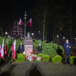 Mężczyzna przemawia podczas uroczystości przy pomniku w nocy. Flagami Polski otoczone są zielone krzewy. Tło to park i oświetlenie.