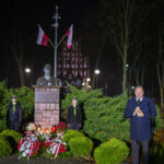 Pomnik otoczony zielonymi krzewami, z flagami Polski nad popiersiem. Widoczne są wiązanki kwiatów w kolorach białym i czerwonym. Na pierwszym planie stoi mężczyzna przy mikrofonie.