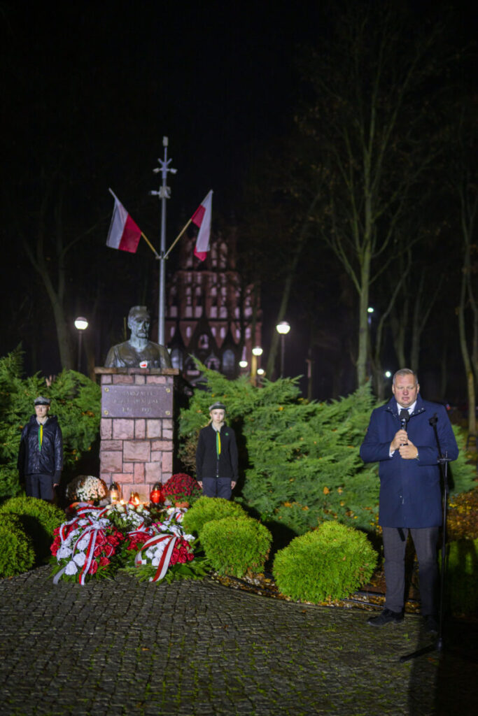 Pomnik otoczony zielonymi krzewami, z flagami Polski nad popiersiem. Widoczne są wiązanki kwiatów w kolorach białym i czerwonym. Na pierwszym planie stoi mężczyzna przy mikrofonie.