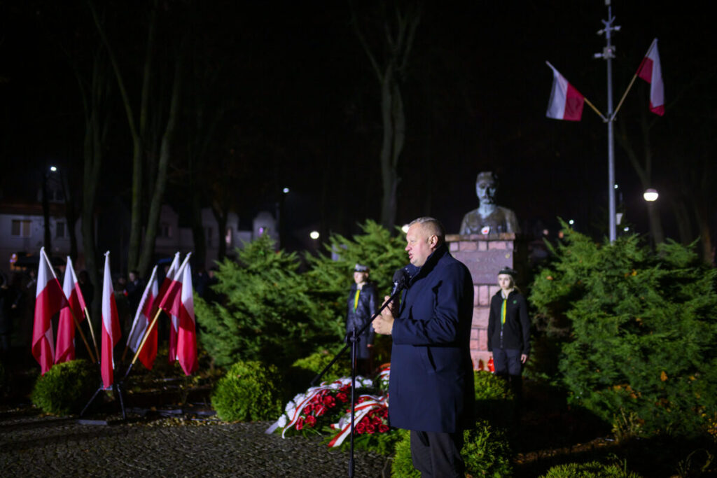 W nocy, mężczyzna przemawia przy mikrofonie obok pomnika. W tle zielone krzewy i polskie flagi. Jasne oświetlenie i ciemne tło dodają kontrastu.