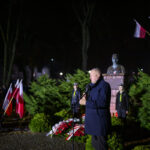 W nocy, mężczyzna przemawia przy mikrofonie obok pomnika. W tle zielone krzewy i polskie flagi. Jasne oświetlenie i ciemne tło dodają kontrastu.