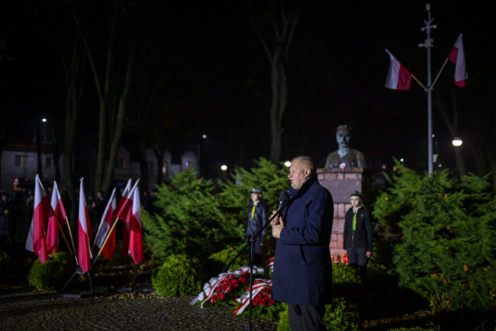 Ujęcie z uroczystości na zewnątrz nocą. Mężczyzna w ciemnym płaszczu mówi do mikrofonu. Wokół polskie flagi i pomnik.