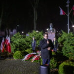 Mężczyzna przemawia na tle zielonych krzewów nocą. Wokół niego stoją flagi Polski oraz wiązanki kwiatów. Za nim znajduje się pomnik i wartownicy.