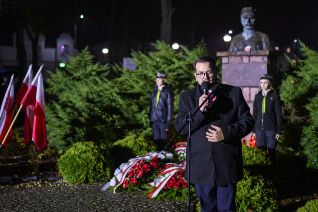 Na zdjęciu mężczyzna przemawia przy mikrofonie, obok flagi Polski. W tle pomnik i zielone krzewy. Nocna atmosfera z oświetleniem ulicznym.
