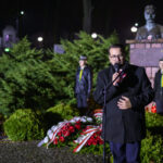 Na zdjęciu mężczyzna przemawia przy mikrofonie, obok flagi Polski. W tle pomnik i zielone krzewy. Nocna atmosfera z oświetleniem ulicznym.