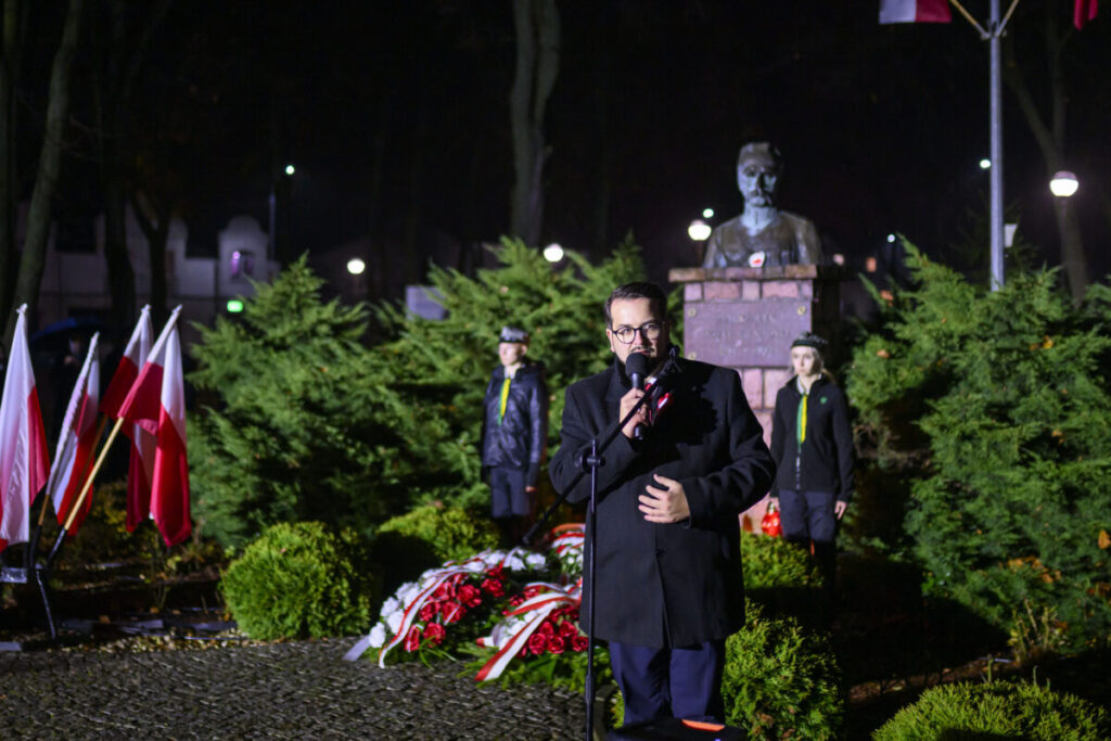Mężczyzna w ciemnym płaszczu przemawia przed mikrofonem na tle zielonych krzewów i popiersia. Widać biało-czerwone flagi i kwiaty. Nocna scena z latarniami w tle.