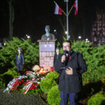Mężczyzna przemawia przy mikrofonie wieczorem, w tle pomnik z popiersiem, flagi i zielone krzewy. Uroczystość z kwiatami.