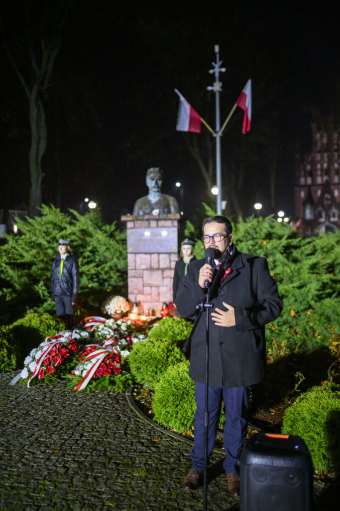 Mężczyzna przemawia przy mikrofonie wieczorem, w tle pomnik z popiersiem, flagi i zielone krzewy. Uroczystość z kwiatami.