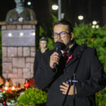 Osoba w okularach przemawia do mikrofonu w parku nocą. W tle popiersie na cokole i zielone krzewy. Oświetlenie tworzy ciepły nastrój.