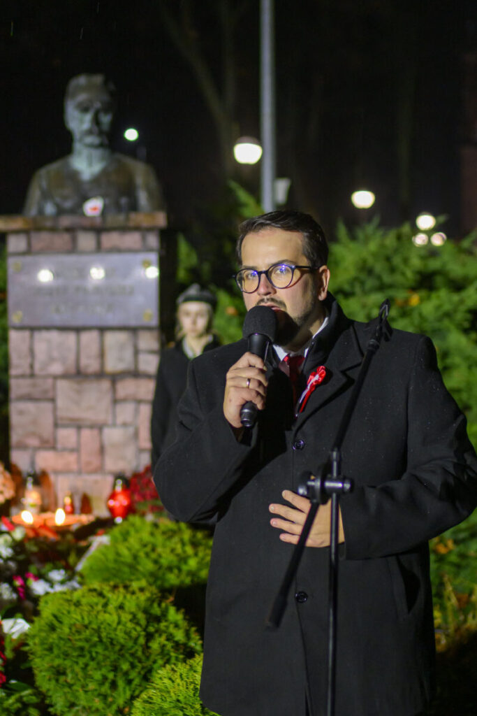 Osoba w okularach przemawia do mikrofonu w parku nocą. W tle popiersie na cokole i zielone krzewy. Oświetlenie tworzy ciepły nastrój.