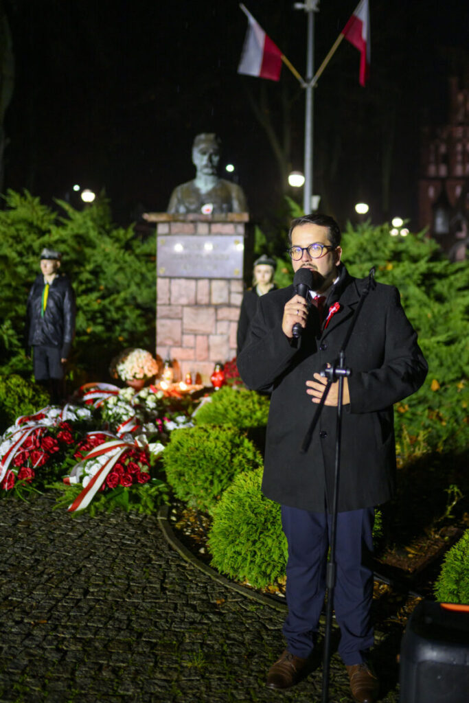 Mężczyzna przemawia do mikrofonu na tle pomnika i flag polskich. Tło to zielone krzewy, kwiaty i znicze. Wieczorna sceneria.