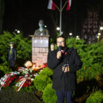 Mężczyzna przemawia przy mikrofonie, obok pomnik z brązu i kwiaty, w tle polskie flagi na maszcie. Nocne oświetlenie rozjaśnia scenę. Ciemne tło z niebieskimi i czerwonymi akcentami.