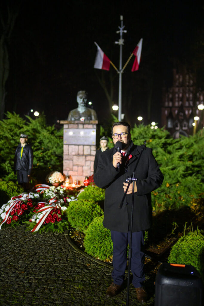 Mężczyzna przemawia przy mikrofonie, obok pomnik z brązu i kwiaty, w tle polskie flagi na maszcie. Nocne oświetlenie rozjaśnia scenę. Ciemne tło z niebieskimi i czerwonymi akcentami.