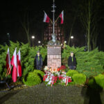 Pomnik otoczony flagami polskimi, noc, oświetlony. Dwóch ludzi w mundurach stoi obok. Świece i kwiaty przed pomnikiem.