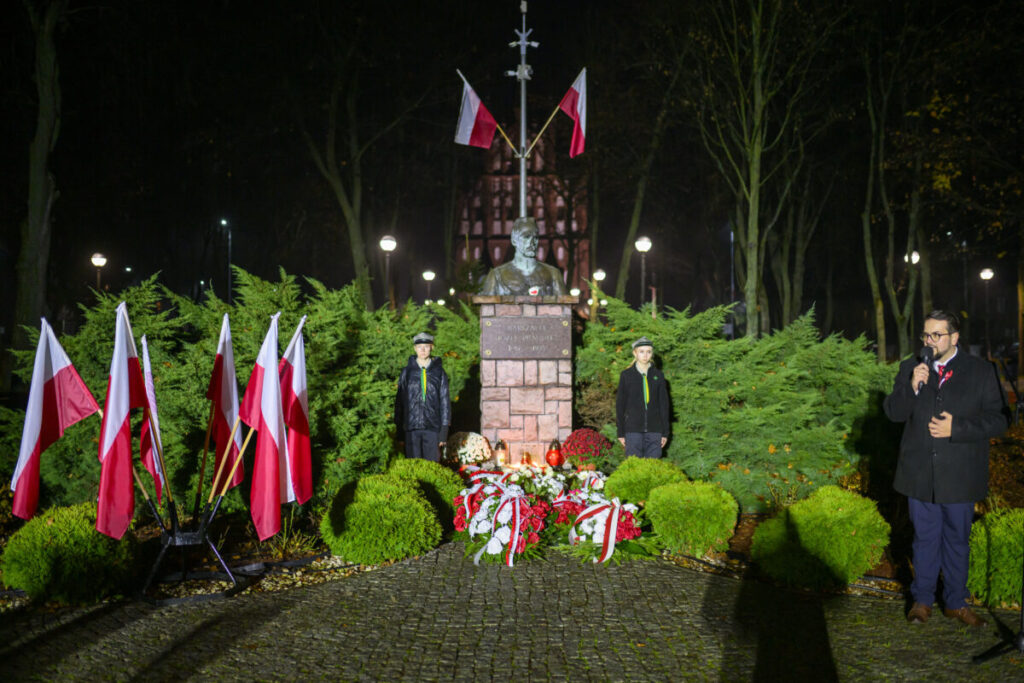 Pomnik z popiersiem, otoczony polskimi flagami, ustawiony na tle zielonych krzewów i oświetlony nocą. Dwie osoby stoją obok monumentu, a trzecia przemawia do mikrofonu. Na pomniku widoczne są wieńce kwiatów.