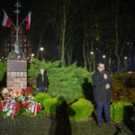 Pomnik z popiersiem i kwiatami, otoczony zielenią, w nocy. Mężczyzna przemawia do mikrofonu. W tle drzewa i światła.