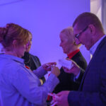 Cztery osoby ściskają wstążki na przyjęciu. Dominują niebieskie światło i półmrok. W tle widać stół z kwiatami i świecami.