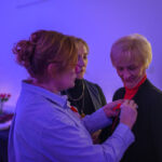 Trzy kobiety w pomieszczeniu z ciepłym, fioletowym oświetleniem. Jedna przypina coś do ubrania innej. W tle stół z jedzeniem i świecami.