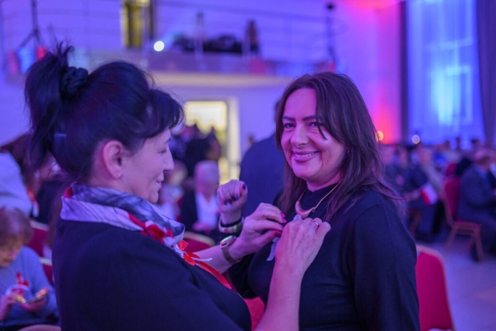 Dwie kobiety w sali z fioletowym oświetleniem, jedna przypina ozdobę drugiej. Tło z rozmytymi sylwetkami ludzi. Czerwone akcenty.