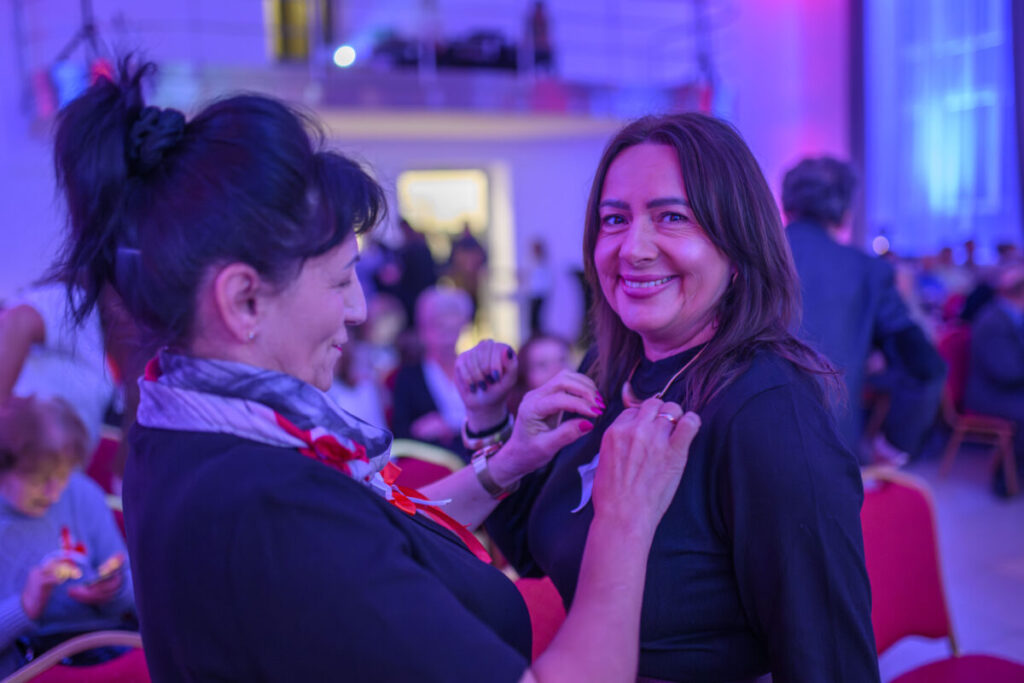 Na zdjęciu dwie kobiety uśmiechają się w sali z fioletowym oświetleniem. Jedna przypina kokardkę do bluzki drugiej. W tle widać ludzi na czerwonych krzesłach.