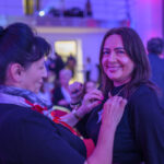 Na zdjęciu dwie kobiety uśmiechają się w sali z fioletowym oświetleniem. Jedna przypina kokardkę do bluzki drugiej. W tle widać ludzi na czerwonych krzesłach.