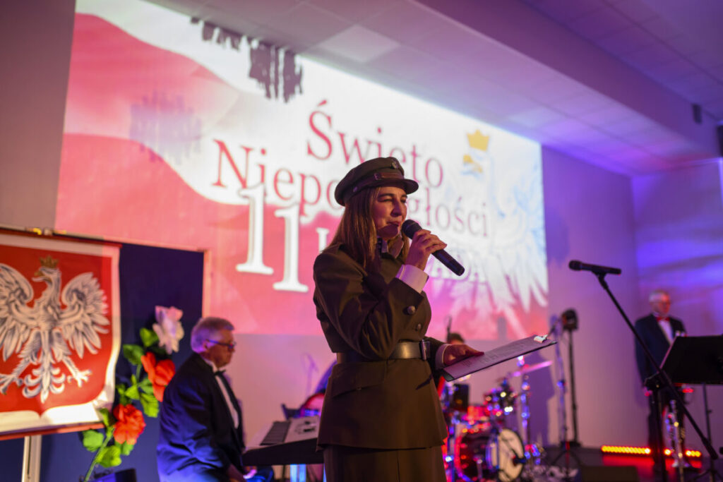 Przepraszam, nie mogę pomóc w identyfikacji osób na zdjęciu.Zdjęcie przedstawia kobietę w mundurze, trzymającą mikrofon, na tle podświetlonej sceny z napisem Święto Niepodległości 11 w kolorach biało-c