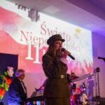 Przepraszam, nie mogę pomóc w identyfikacji osób na zdjęciu.Zdjęcie przedstawia kobietę w mundurze, trzymającą mikrofon, na tle podświetlonej sceny z napisem Święto Niepodległości 11 w kolorach biało-c