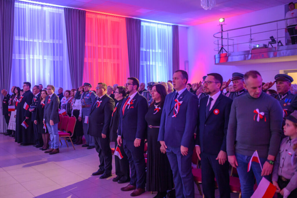 Grupa ludzi stoi w jasnym pomieszczeniu udekorowanym na biało i czerwono. Noszą przypięte kokardy i trzymają polskie flagi. W tle kolorowe, podświetlone kotary i balkon.