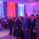 Grupa ludzi stoi w jasnym pomieszczeniu udekorowanym na biało i czerwono. Noszą przypięte kokardy i trzymają polskie flagi. W tle kolorowe, podświetlone kotary i balkon.