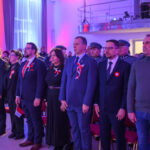 Uroczystość z udziałem osób w garniturach i mundurach. Trzymają polskie flagi, w tle czerwono-niebieskie światła. Sala pełna uczestników, patriotyczny nastrój.