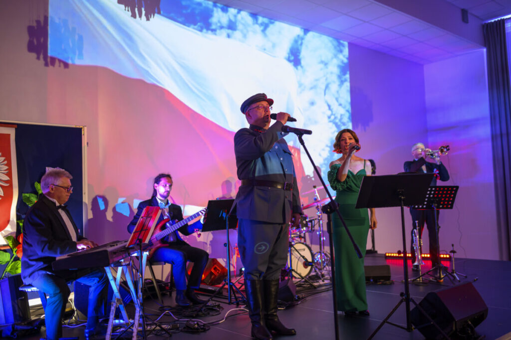 Grupa muzyków występuje na scenie z projekcją polskiej flagi w tle. Na pierwszym planie śpiewają mężczyzna i kobieta. Kolorowy wystrój podkreśla uroczystość wydarzenia.